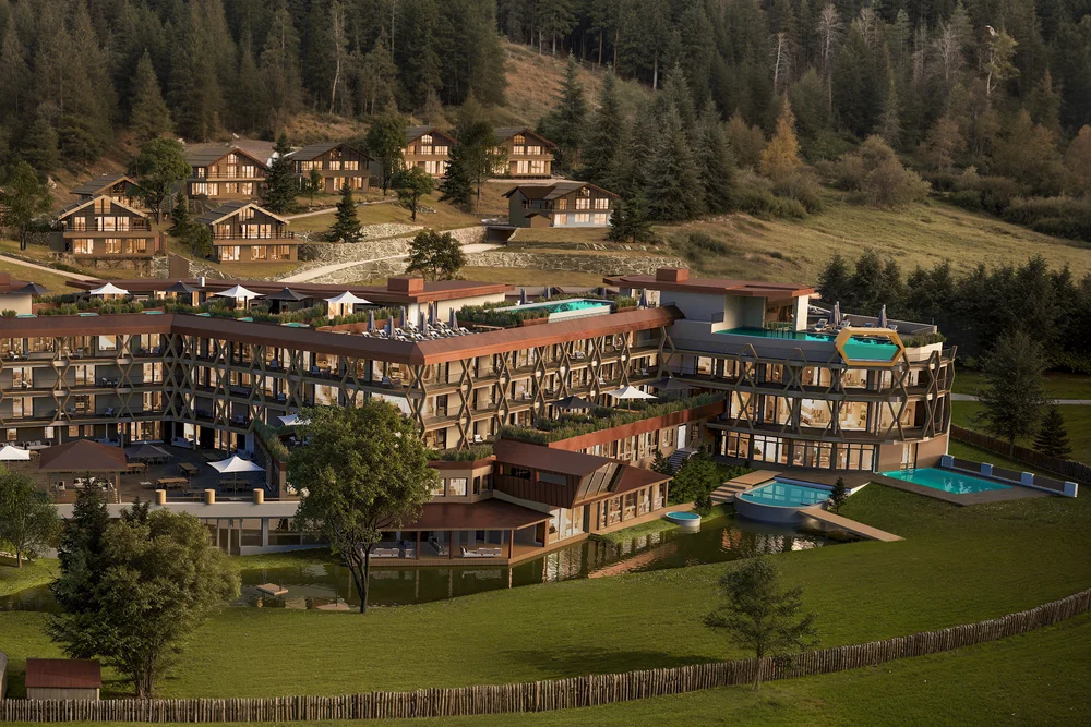 Aerial view of Alpura in summer: hexagonal main building with pools on staggered rooftop terraces, set within dense pine forest. Chalets on the hillside in the background, playground and pond on the meadow in the foreground.