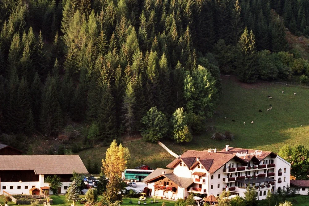 Bird's-eye view of the Alphotel Tyrol in its historic farmstead setting: main building, barn and pond nestled between pastures and mountain forest — the origin of today's Alpura.